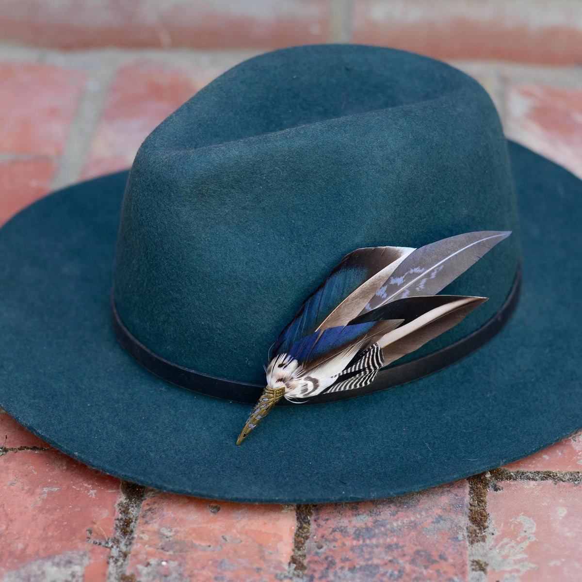 A iridescent blue mallard duck wing feather hat pin  on a teal fedora resting on a brick surface