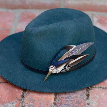 A iridescent blue mallard duck wing feather hat pin  on a teal fedora resting on a brick surface