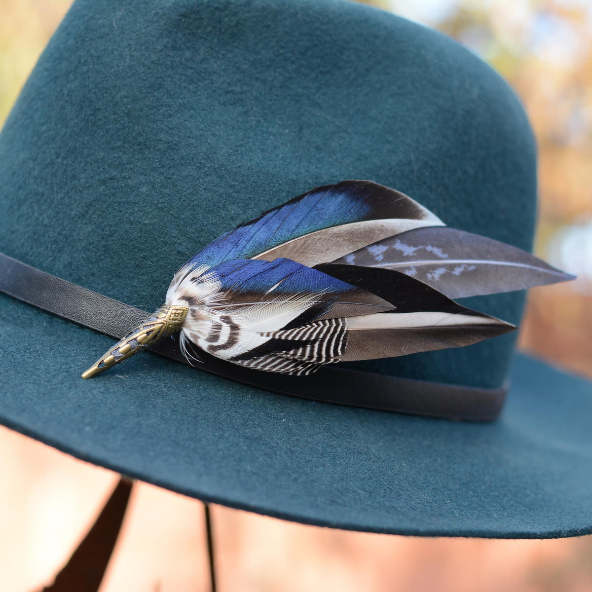 A close-up of a teal wide-brim hat with a black band and a decorative duck feather hat pin in blue, black, white, and brown tones.