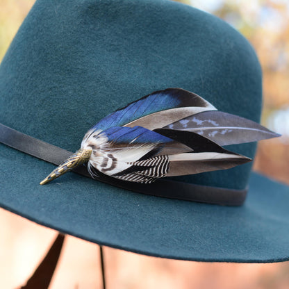 A close-up of a teal wide-brim hat with a black band and a decorative duck feather hat pin in blue, black, white, and brown tones.