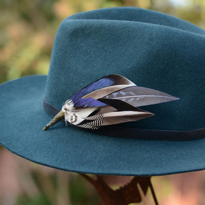 A dark teal felt wide-brim hat with a black band and a duck feather hat pin in iridescent blue, black and grey, photographed outdoors against a blurred natural background.