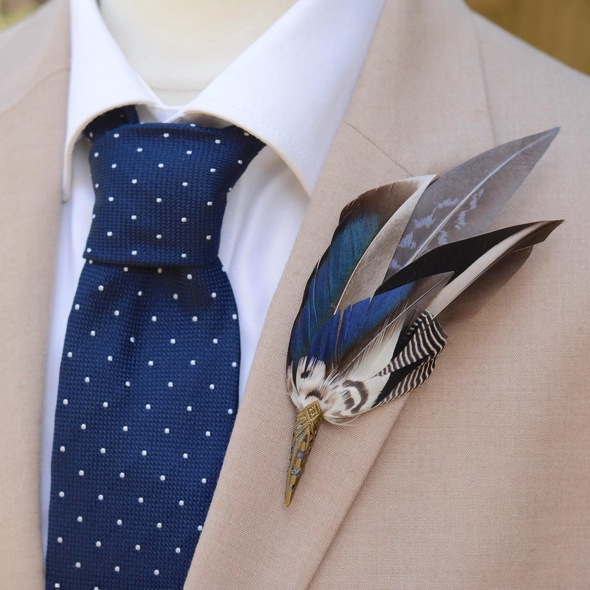 A close-up of a men's formal outfit featuring a navy blue tie with white polka dots, a white dress shirt, and a beige suit jacket. A decorative feathered boutonniere is pinned to the left lapel. The boutonniere features an elegant sweep of duck feathers in iridescent blue and graphic black and white markings.