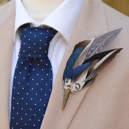 A close-up of a men's formal outfit featuring a navy blue tie with white polka dots, a white dress shirt, and a beige suit jacket. A decorative feathered boutonniere is pinned to the left lapel. The boutonniere features an elegant sweep of duck feathers in iridescent blue and graphic black and white markings.