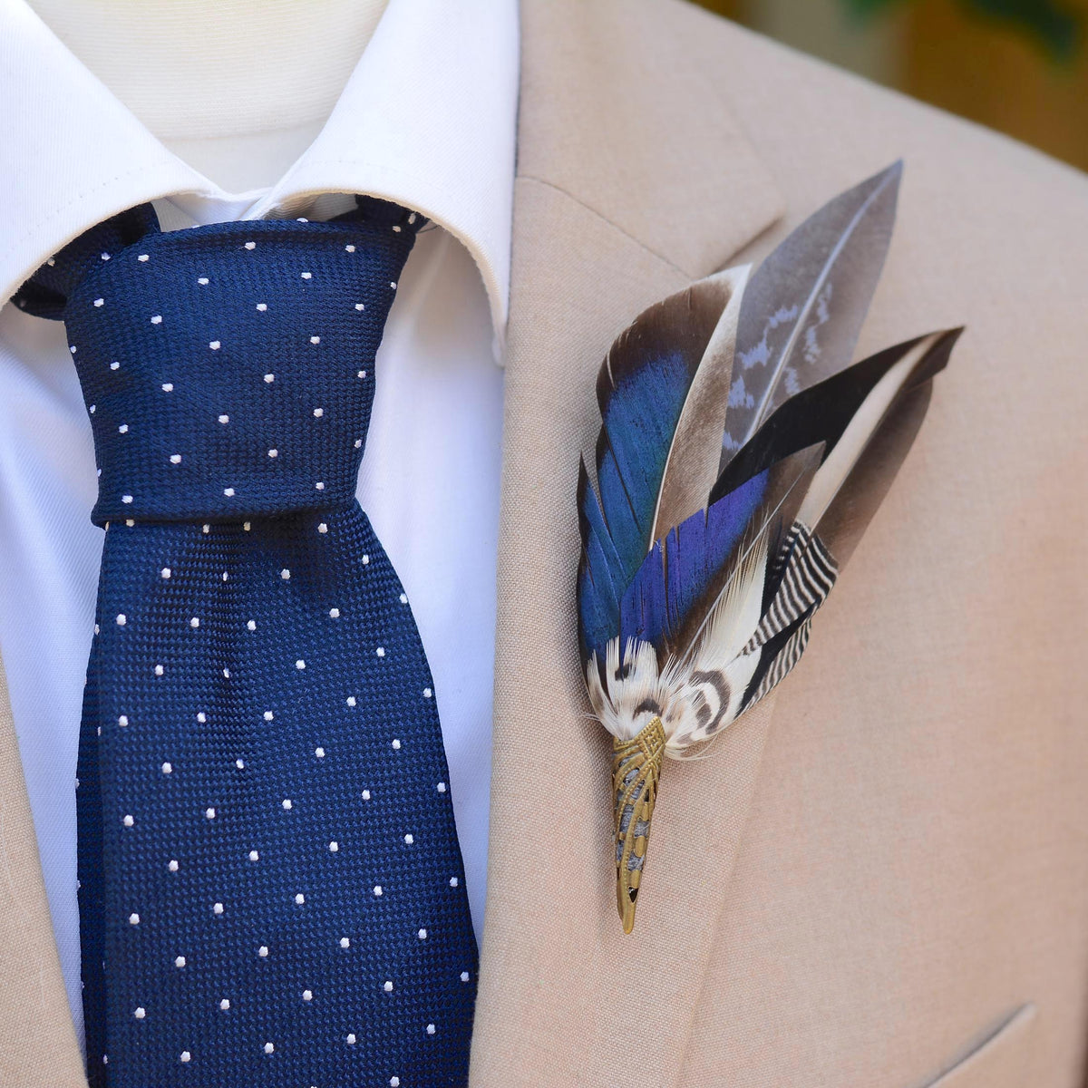 A close-up of a men's formal outfit featuring a navy blue tie with white polka dots, a white dress shirt, and a beige suit jacket. A decorative feathered boutonniere is pinned to the left lapel. The boutonniere features an elegant sweep of duck feathers in iridescent blue and graphic black and white markings.