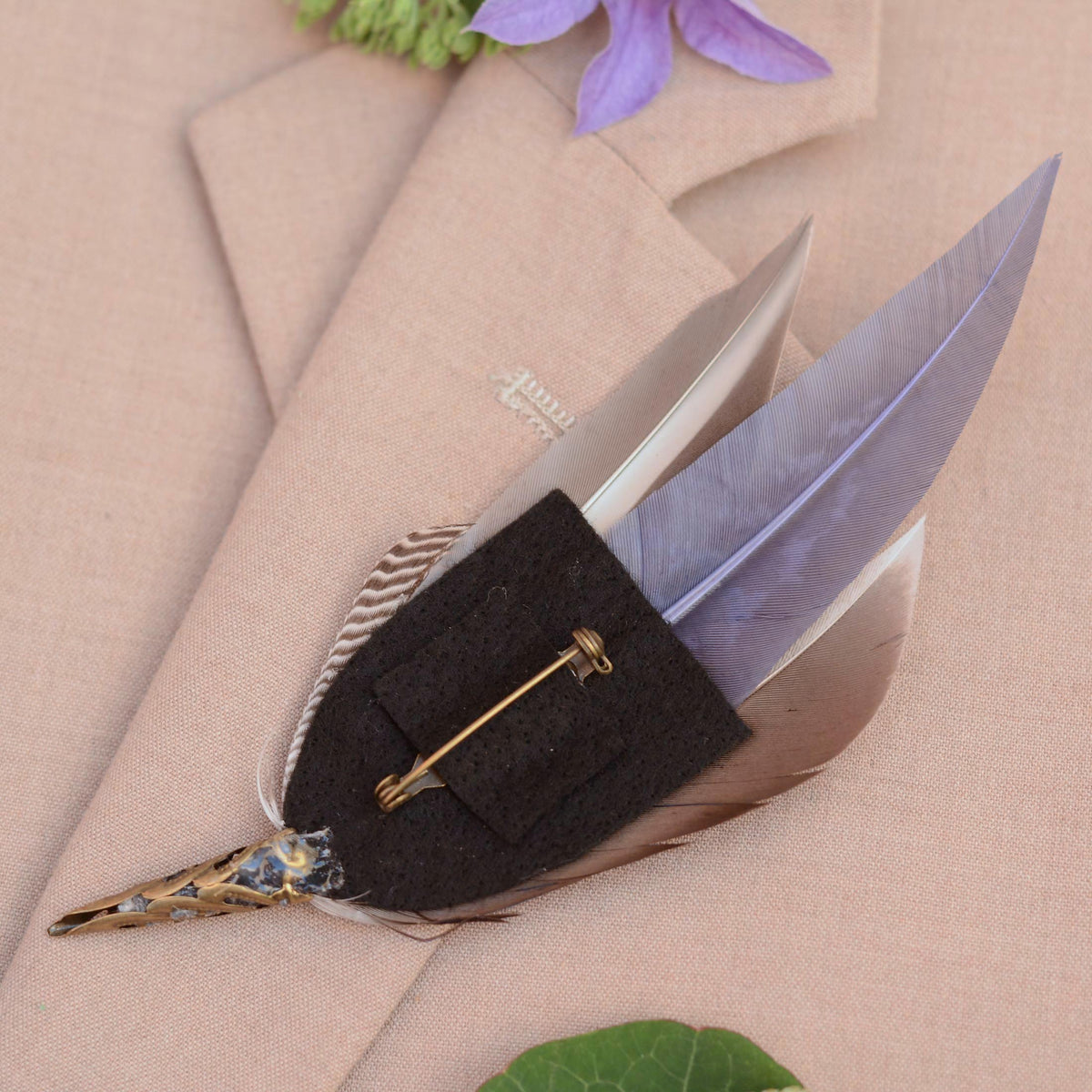 A decorative feather pin with a black felt base, and brass-toned metal pin, displayed on a beige textured fabric background.
