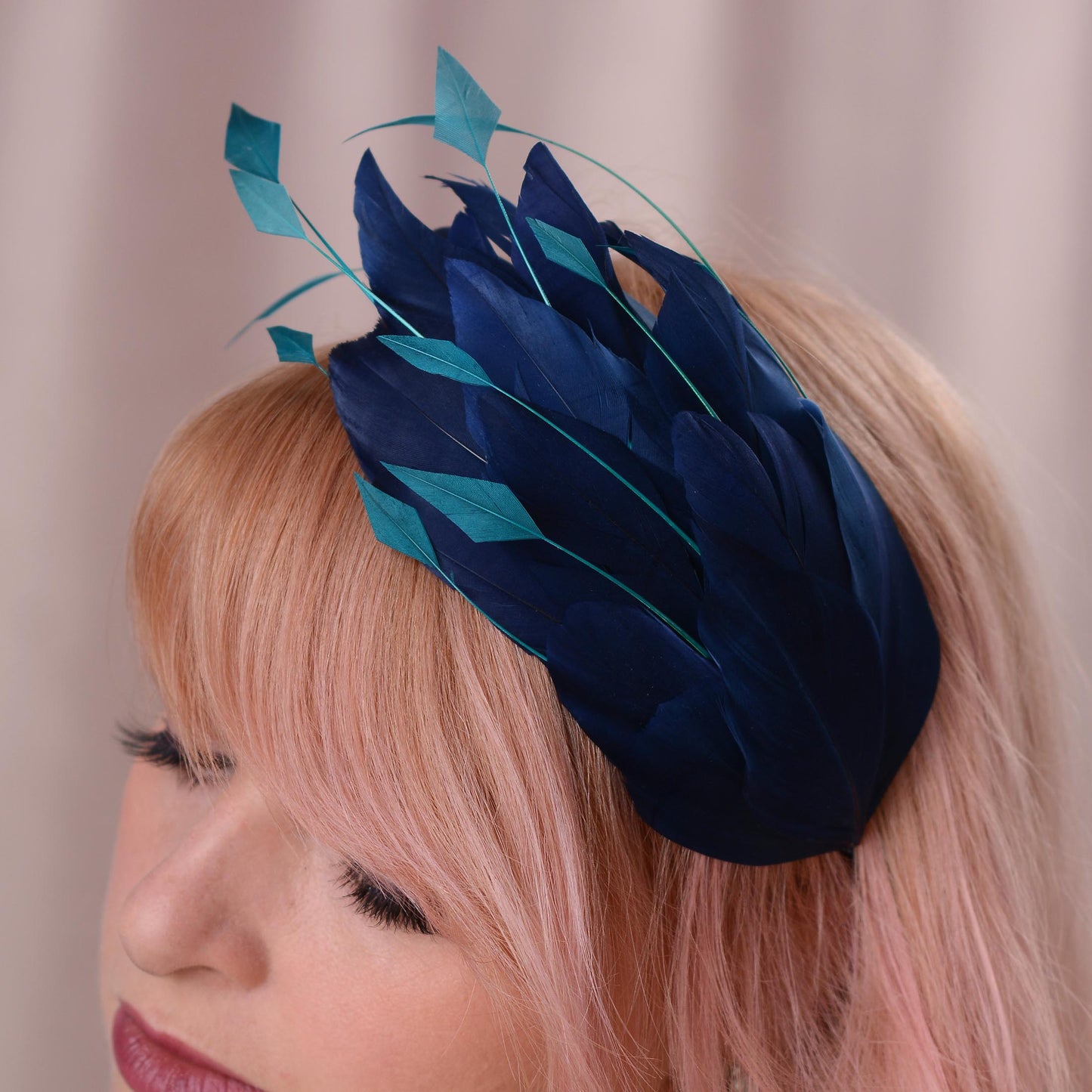 A close-up of a woman with pink hair wearing a decorative fascinator headpiece made of layered feathers in dark navy and teal.