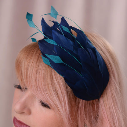 A close-up of a woman with pink hair wearing a decorative fascinator headpiece made of layered feathers in dark navy and teal.