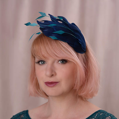 A woman with short pink hair wearing a navy blue  and teal goose feather fascinator headpiece, photographed against a soft beige curtain background.