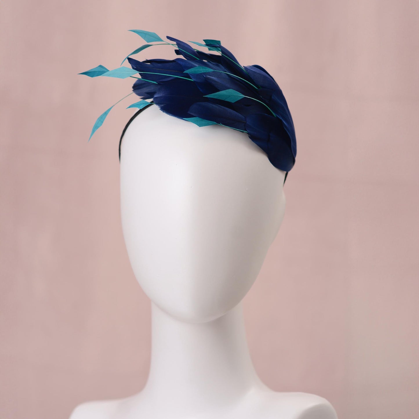 A navy blue feather wedding fascinator headpiece with teal accents, displayed on a white mannequin head against a soft pink background.