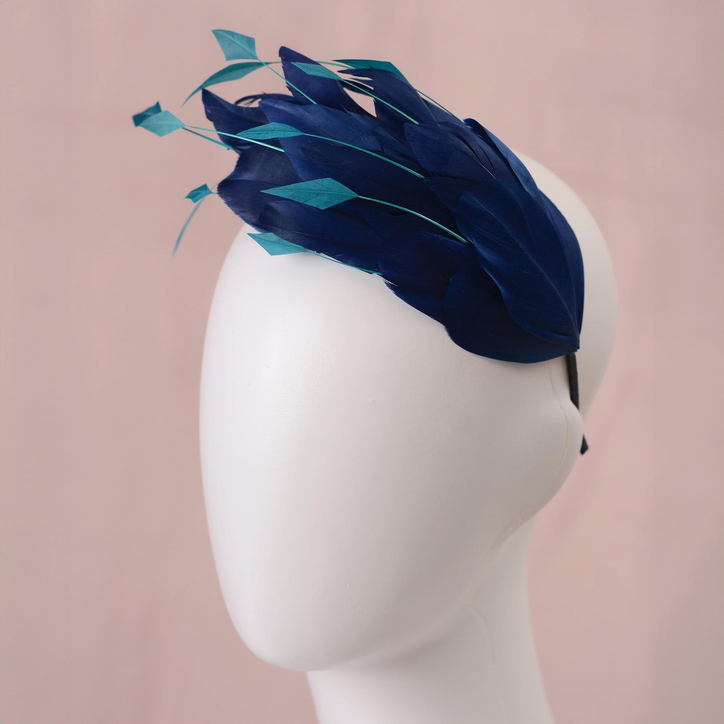 A navy blue and teal formal event fascinator feather headpiece displayed on a white mannequin head against a soft pink background.