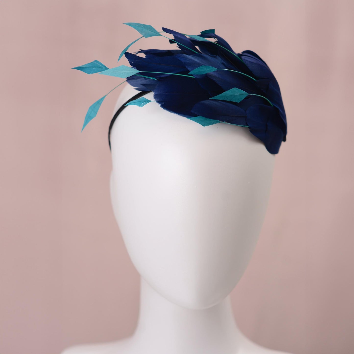 A navy blue feather mother of the bride fascinator headpiece with turquoise teal accents, displayed on a white mannequin head against a soft pink background.