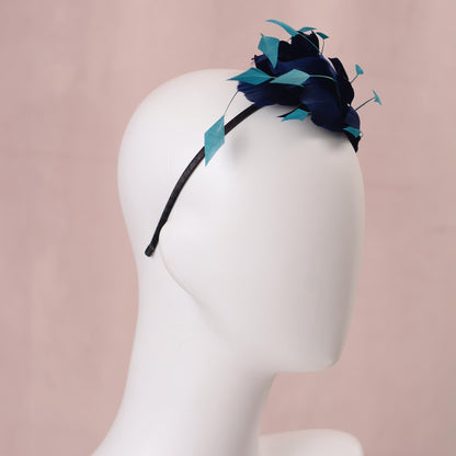 A navy blue feather side perching fascinator headpiece with turquoise teal accents, displayed on a white mannequin head against a soft pink background.