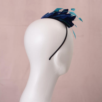 A navy blue feather side perching fascinator headpiece with floating turquoise teal accents, displayed on a white mannequin head against a soft pink background.