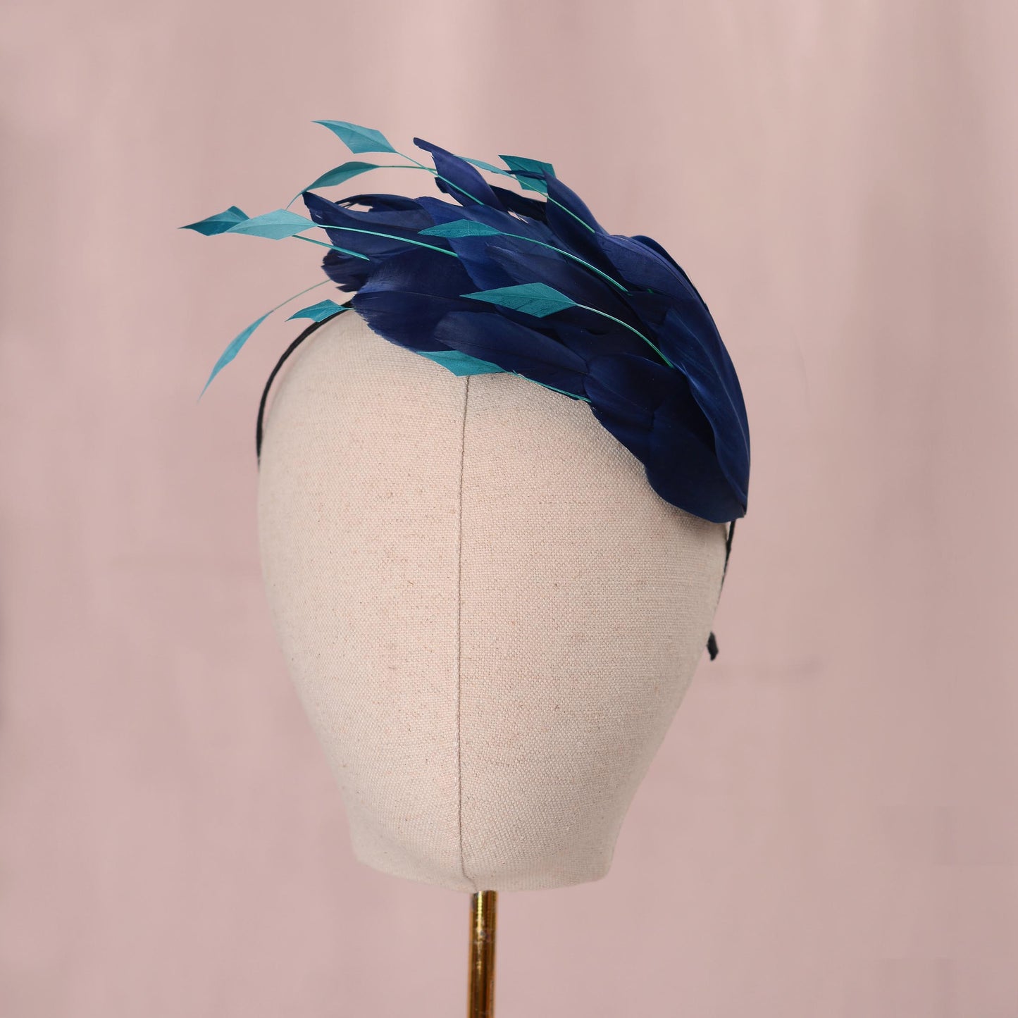 A party fascinator headpiece with dark navy and teal feathers, displayed on a cream fabric mannequin head against a soft pink background.
