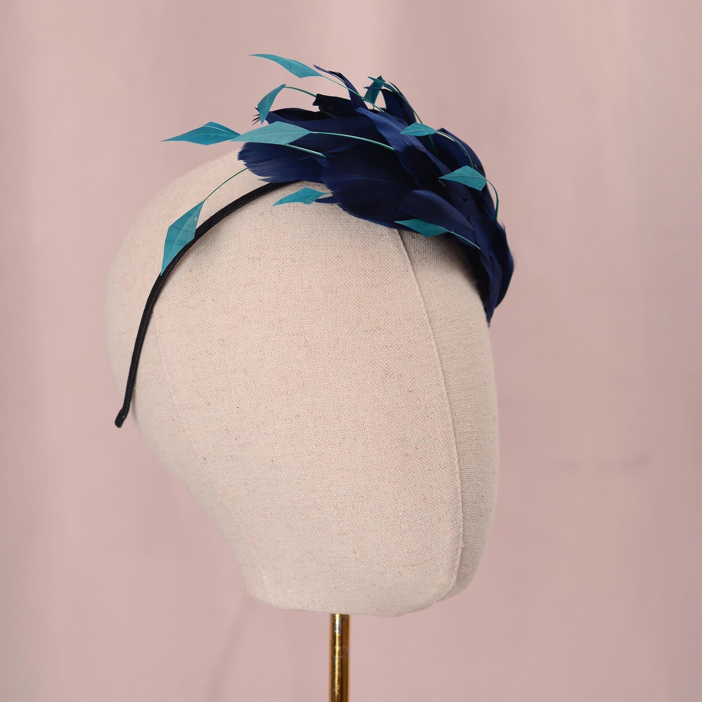 A party fascinator headpiece with dark navy and teal feathers, displayed on a cream fabric mannequin head against a soft pink background.