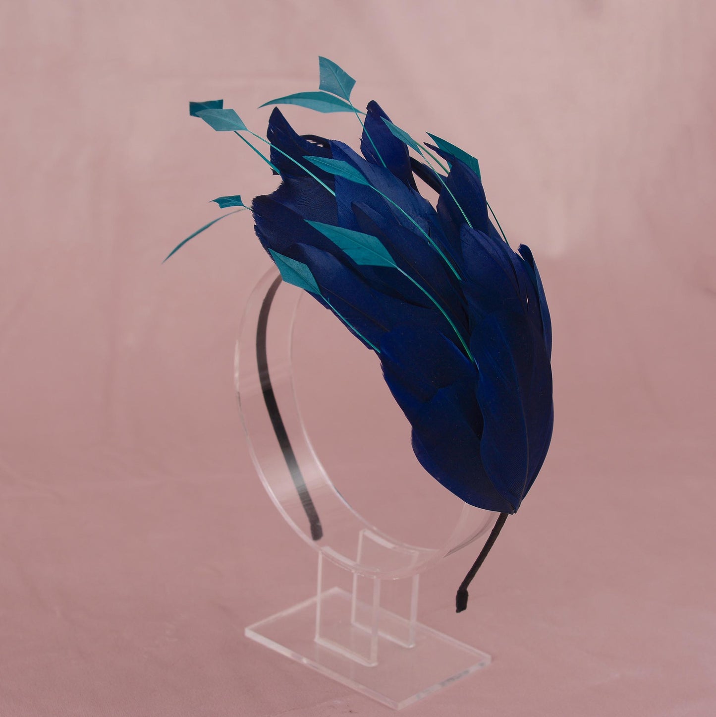 A blue and teal feather fascinator headpiece displayed on a clear acrylic stand against a dusty pink background.