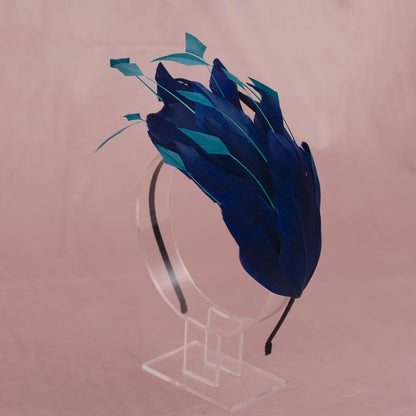 A blue and teal feather fascinator headpiece displayed on a clear acrylic stand against a dusty pink background.