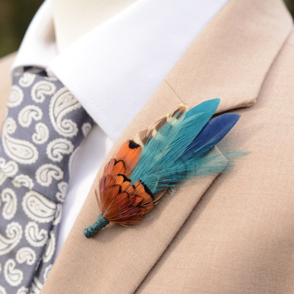 Feathered boutonniere on a beige suit jacket with a patterned tie. The buttonhole is crafted with copper pheasant feathers paired with teal and navy.