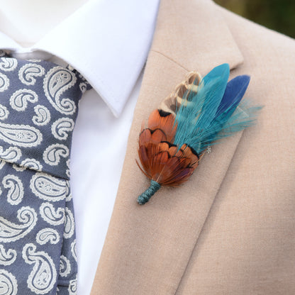 Feathered wedding boutonniere on a suit lapel with a patterned tie. Featuring copper pheasant feathers paired with teal and navy perfect for countryside weddings.