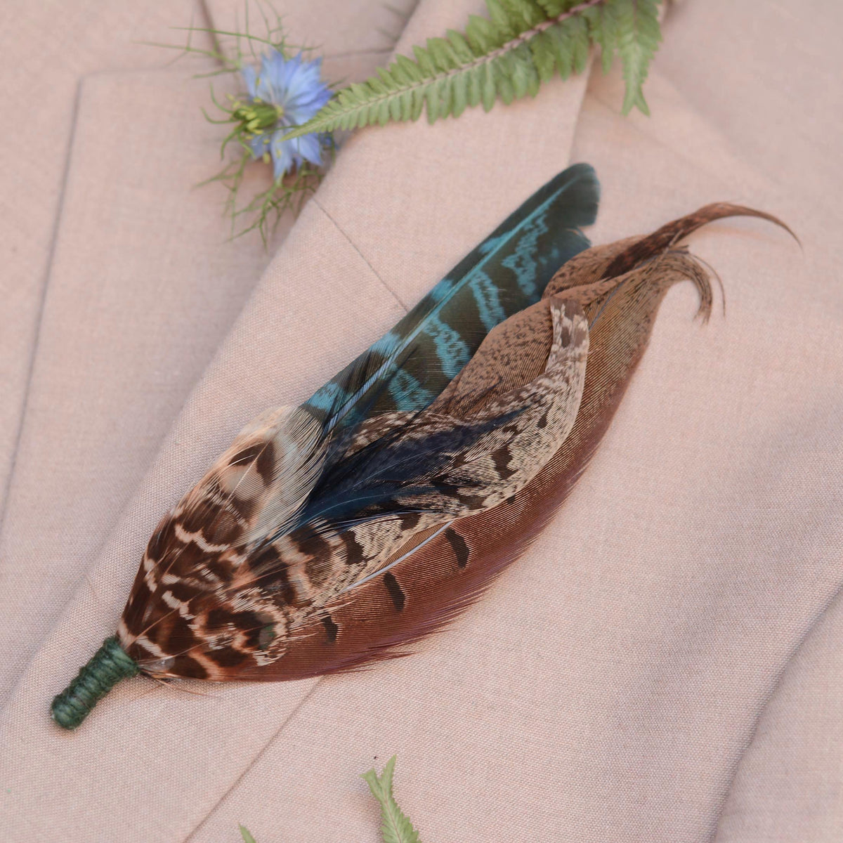 A decorative feather pin featuring a brown patterned duck feather with teal accents, and a green twine wrapped stem, displayed on a beige fabric background with a small blue flower and green leaves.