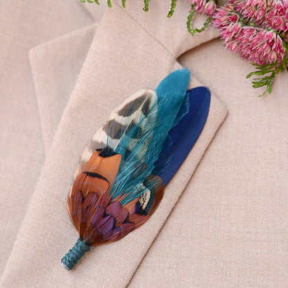 Colourful pheasant feather brooch in copper, teal and navy on a beige suit lapel background with pink flowers.