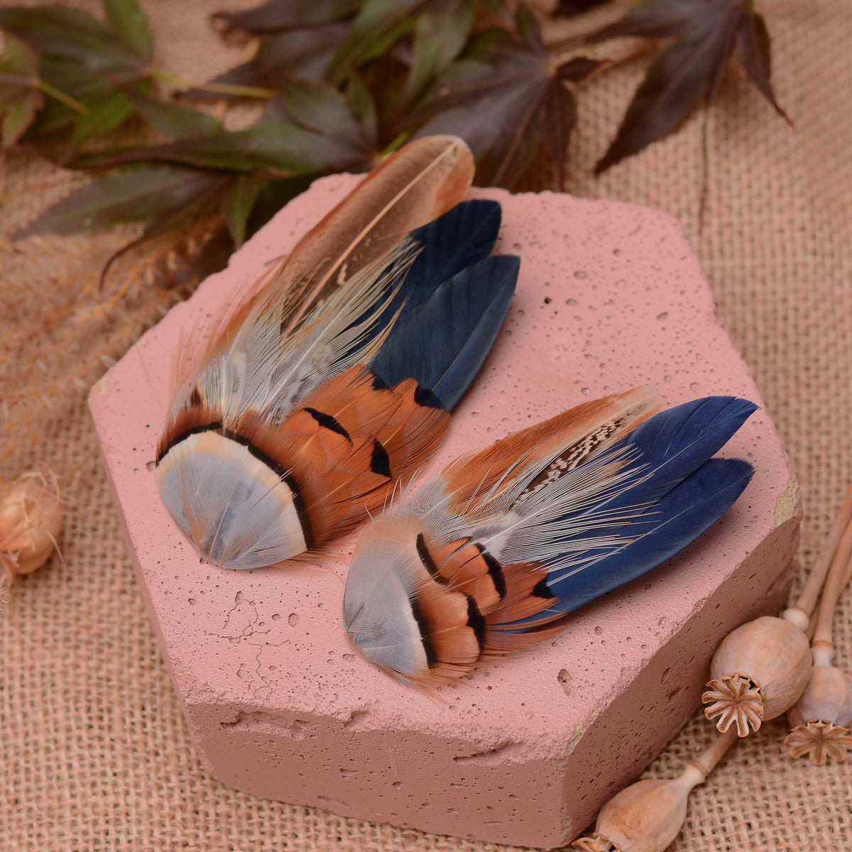 Two decorative bird feathers fascinator hair clip with blue, brown, coper and black markings, arranged on a pink stone slab with burlap and dried leaves in the background.
