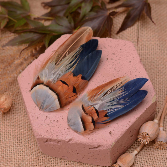 Two decorative bird feathers fascinator hair clip with blue, brown, coper and black markings, arranged on a pink stone slab with burlap and dried leaves in the background.