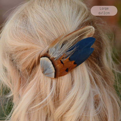 A large feather hair accessory clipped onto blonde hair, featuring a blue feather, a brown feather with black markings, and a pale grey feather.