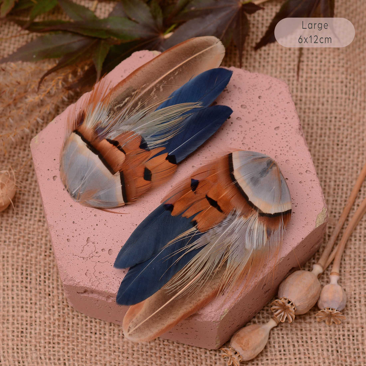 Two decorative large bird feather hair clips, in blue, browns and copper displayed on a pink hexagonal block over a burlap background with dried botanical props. Perfect accent for Navy blue dresses