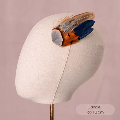 A large feather fascinator accessory displayed on a beige fabric mannequin head. The feather features a mix of brown, orange, blue, and grey feathers.