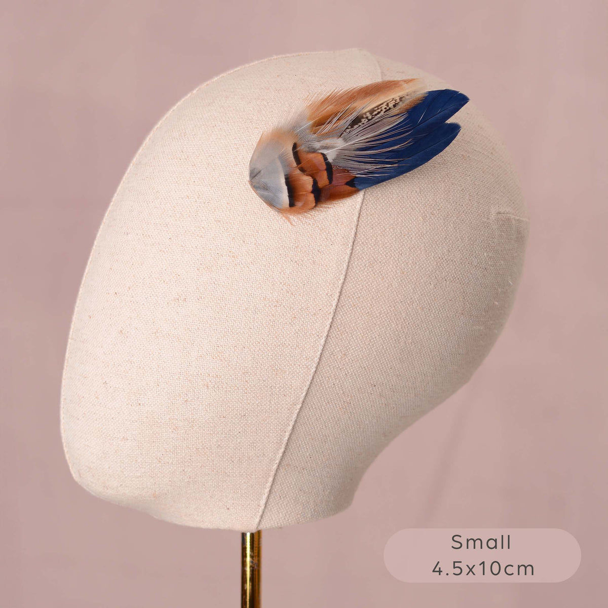 A small decorative feather hair accessory with blue, brown, white, and grey feathers, displayed on a beige fabric mannequin head against a soft pink background. 