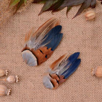Two decorative bird feather hair pieces with blue, brown, tan, and copper feathers, arranged on a beige burlap surface with scattered dried poppy pods and leaves.