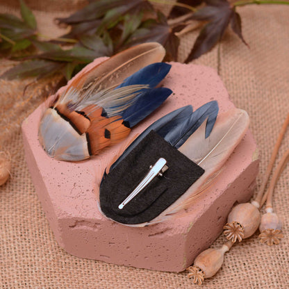 Two decorative feather hair clips displayed on a pink stone block over a burlap surface. One clip features a large beige and brown feather with blue accents, while the other is upside down to show the black felt base and a silver-toned metal alligator clip.