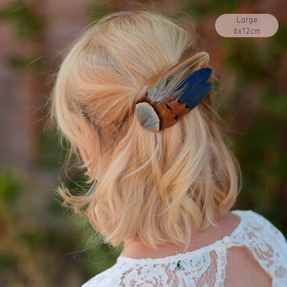 A blonde woman shown from the back wearing a large feather hair clip in a bohemian style. The clip features navy blue, brown, copper and grey blue in a wing shape