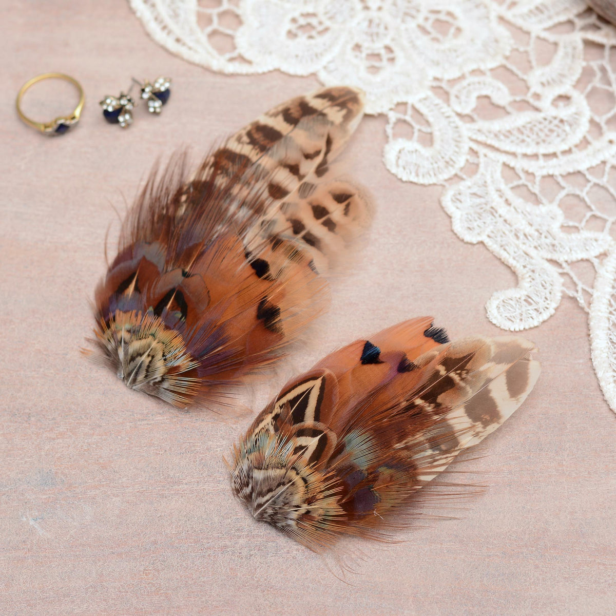 Two decorative pheasant feather hair clip pieces with brown, tan, black, and muted iridescent tones, arranged on a pale pink surface with white lace and small jewellery props nearby.
