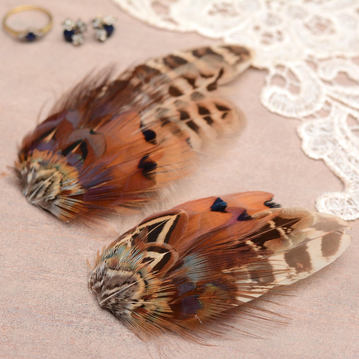 Two decorative feather hair accessories with intricate brown, beige, and black patterned pheasant feathers, arranged on a beige surface with white lace and small jewellery props in the background.