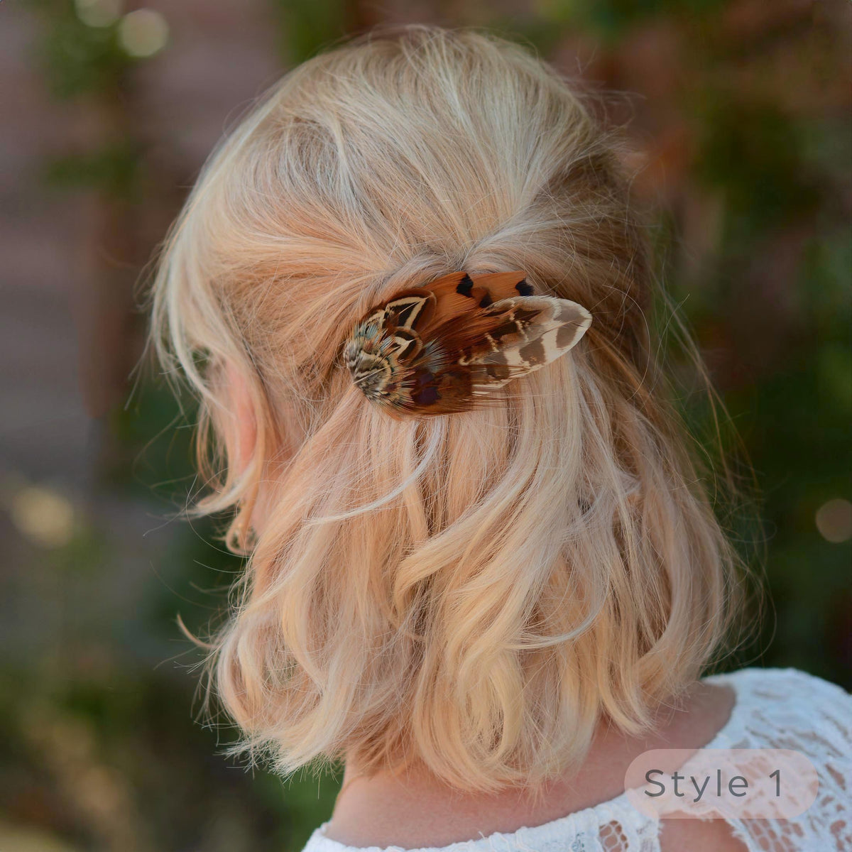 A blonde woman shown from the back  with a bob in a half up do wearing a brown and beige pheasant feather hair clip in her hair, with a soft-focus outdoor background. 