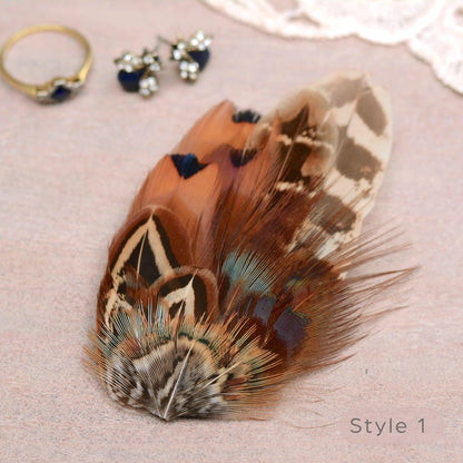 A decorative pheasant feather accessory with intricate brown, beige, black, and blue patterns, styled on a textured pink surface alongside jewellery props.