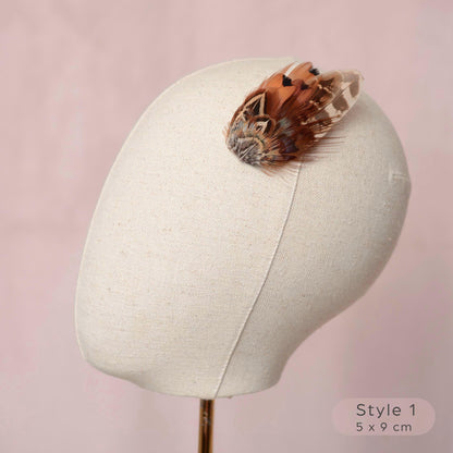 A decorative pheasant feather hair accessory in warm brown, tan, and cream tones, displayed on a cream fabric mannequin head against a soft pink background. 