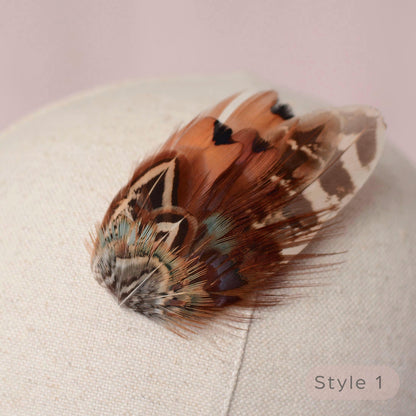 A decorative rustic pheasant feather hair accessory with intricate brown, black, white, and teal patterned feathers, displayed on a textured cream fabric surface.