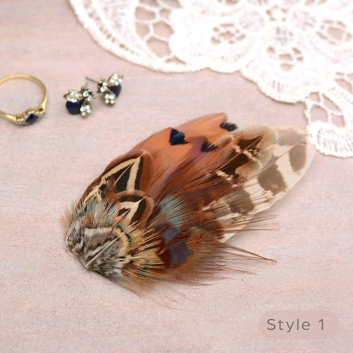 A decorative pheasant feather hair accessory with brown, beige, black, and iridescent blue-green patterning, styled on a light pink surface with lace trim, a gold ring, and small stud earrings nearby. The text 'Style 1' appears in the lower right.