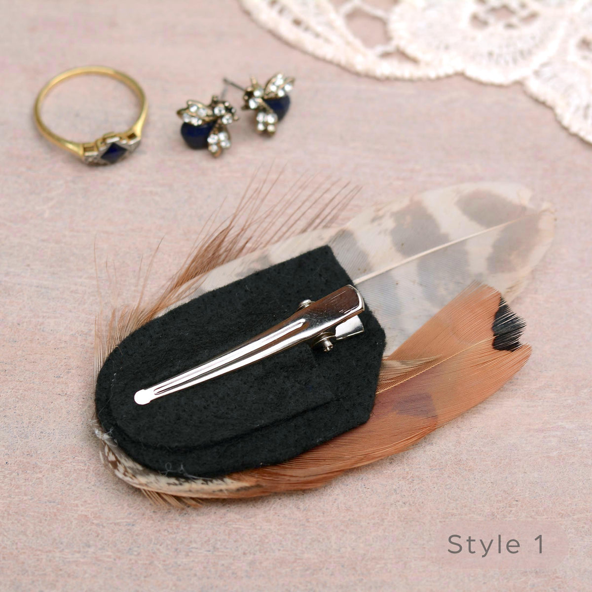 A feather hair clip accessory displayed on a textured pink surface, on a black felt pad, with a silver alligator style metal clip, and feathers in brown, tan, and cream. A gold ring and blue-and-silver stud earrings are placed nearby as styling props,
