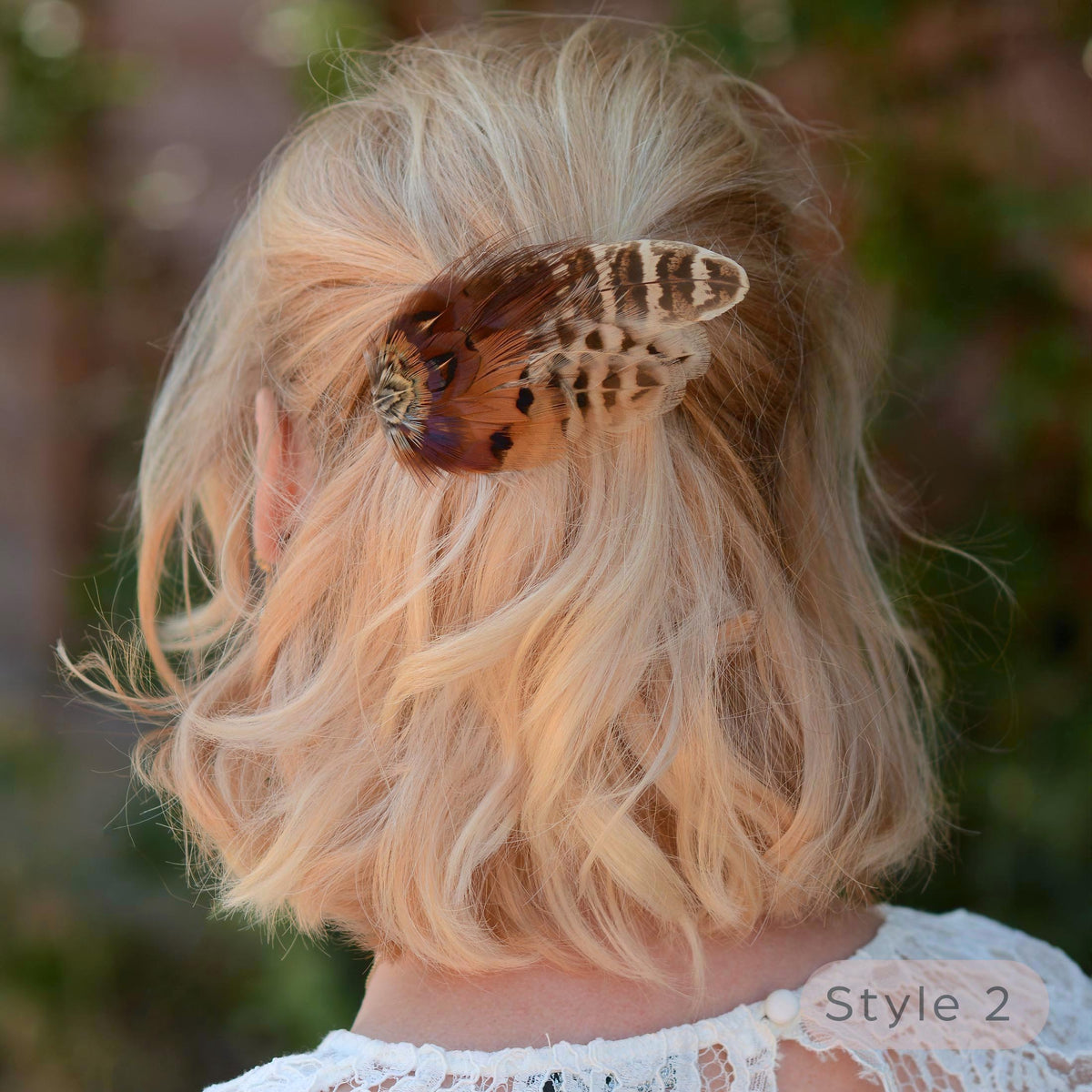 A blonde woman with a bob in a half up do shown from the back wearing a rustic brown and beige pheasant feather hair clip in her hair, with a soft blurred outdoor background.