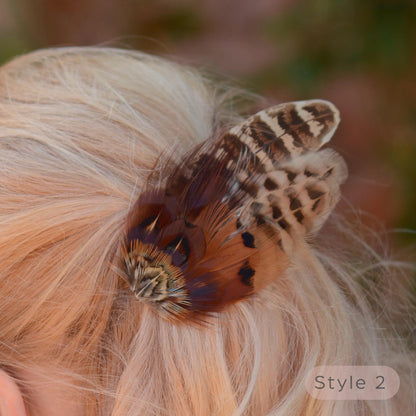 Close up of a blonde woman with a bob in a half up do shown from the back wearing a rustic brown and beige pheasant feather hair clip in her hair, with a soft blurred outdoor background.