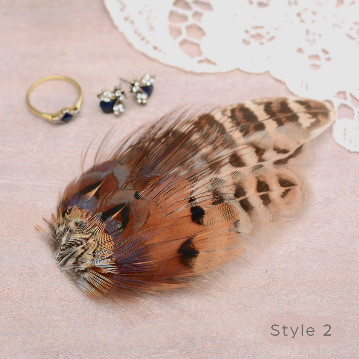 A decorative feather hair accessory with brown, beige, and black patterned plumes arranged on a soft pink surface, styled with small jewellery pieces and lace. Ideal for a rustic or barn wedding and countryside events