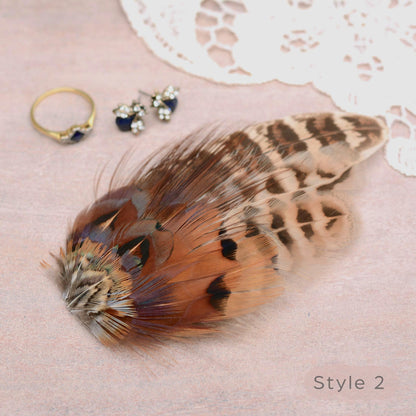 A decorative feather hair accessory with brown, beige, and black patterned plumes arranged on a soft pink surface, styled with small jewellery pieces and lace. Ideal for a rustic or barn wedding and countryside events