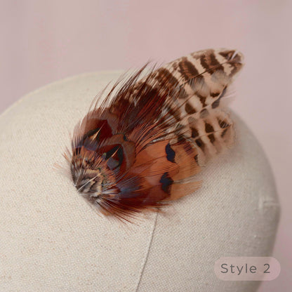 A decorative feather hair accessory with brown, black, and beige patterned feathers, displayed on a cream fabric mannequin head against a soft pink background.