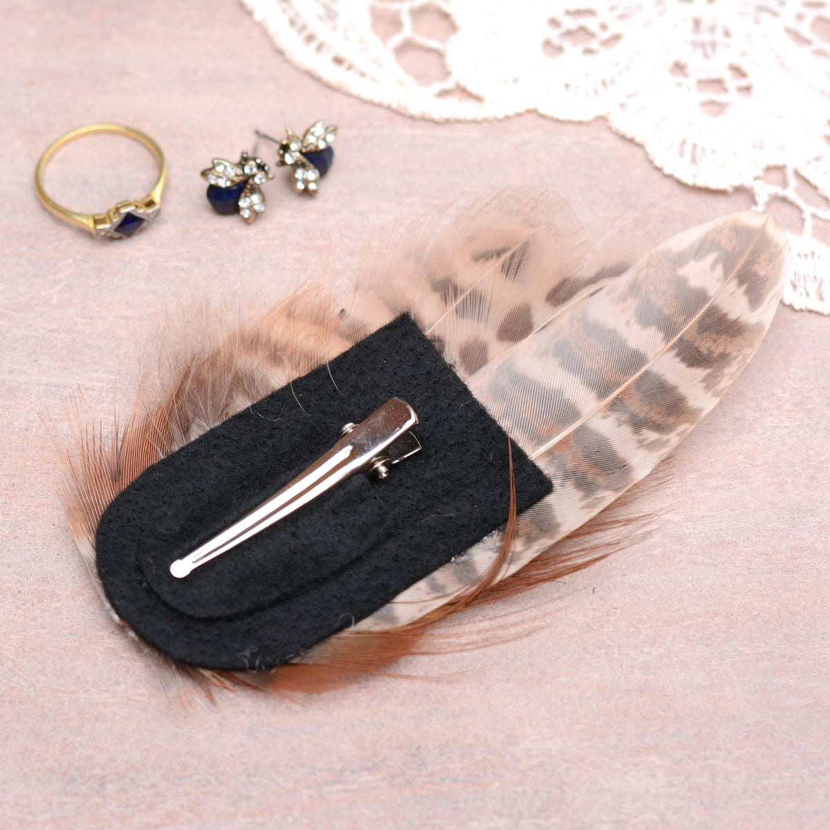 A feather hair clip with a black felt base and silver metal alligator style hairpin, displayed on a textured pink surface with lace and jewellery props.