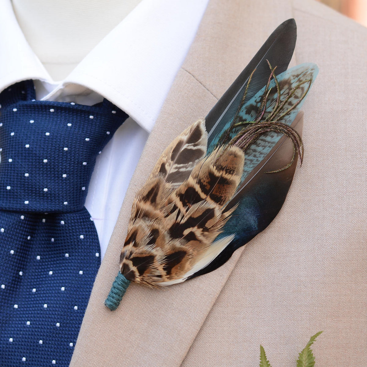 A decorative feather lapel pin with brown, black, and teal feathers, pinned to a beige suit jacket over a white dress shirt and a navy polka dot tie.