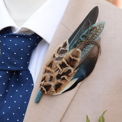 A decorative feather lapel pin with brown, black, and teal feathers, pinned to a beige suit jacket over a white dress shirt and a navy polka dot tie.